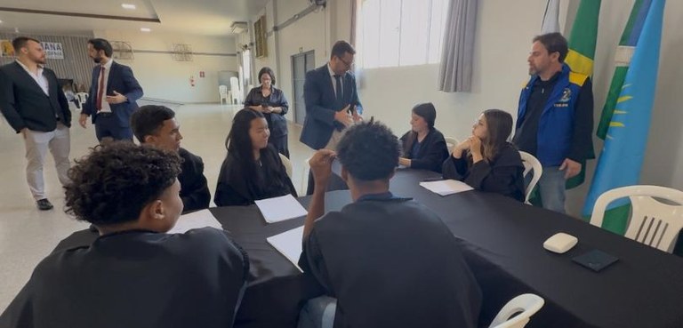 Fotografia em que são vistos vários jovens sentados em torno de uma mesa coberta por uma toalha ...