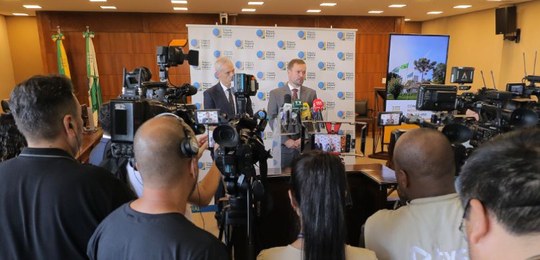 Fotografia que retrata uma coletiva de imprensa em ambiente interno. Em plano intermediário, doi...