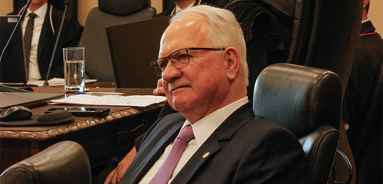 Fotografia que retrata um homem branco idoso, de cabelos brancos e curtos, usando óculos de arma...