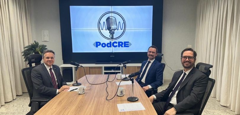 Novo episódio do PodCRE discute atuação do Ministério Público nas Eleições 2024 Fotografia de três homens em cadeiras pretas ao redor de uma mesa de madeira retangular. Acima d...