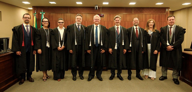 Fotografia de nove pessoas, seis homens e três mulheres, posando em pé lado a lado em um ambient...