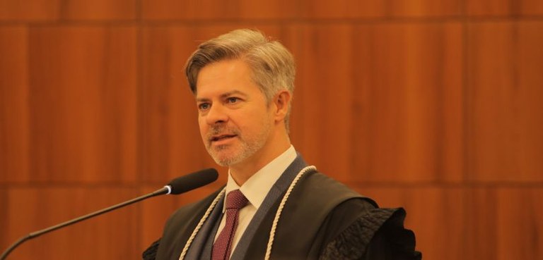 Fotografia de um homem branco com cabelos grisalhos falando diante de um microfone em uma sala c...