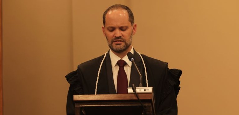 Fotografia de um homem branco, com cabelos castanhos falando diante de um microfone, que aparece...