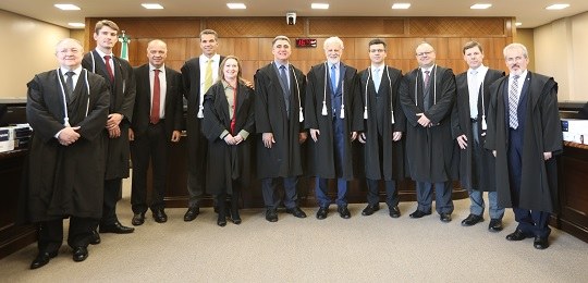 Fotografia de um grupo de juízes e uma juíza em pé em uma sala. Todos usam togas pretas por cima...