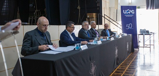 Fotografia de cinco homens sentados em uma mesa, todos eles estão vestindo terno.