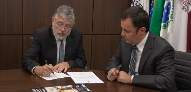 Na foto, dois homens estão sentados em uma mesa. Um deles está assinando um documento enquanto o...