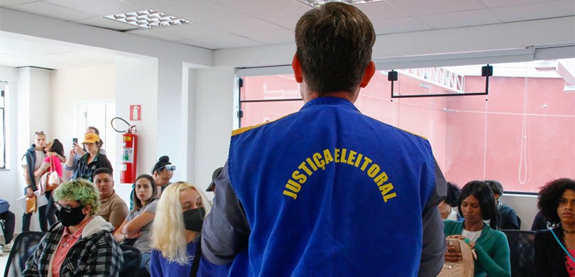 Ouvidoria do TRE-PR participa de mutirão para retificação de prenome e gênero para pessoas trans Fotografia de um homem de costas, vestindo o colete da Justiça Eleitoral. Ele está de frente par...