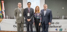 Em frente a uma mesa com um banner branco, três homens e uma mulher posam para a foto.