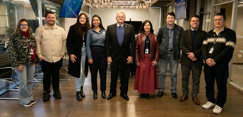 Fotografia em que se veem cinco homens e quatro mulheres posando em pé sem uma sala com piso mar...