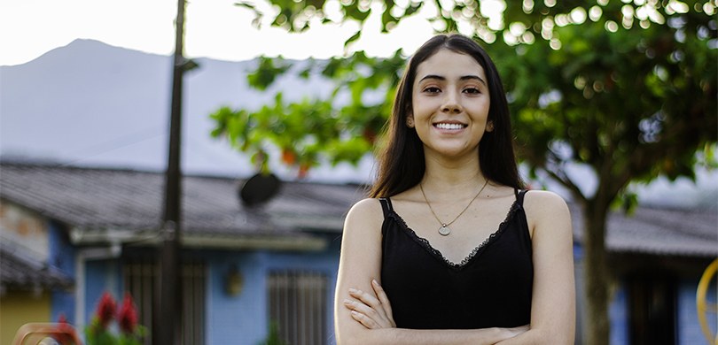 foto em que se vê uma mulher vestindo uma regata preta e um colar dourado, sorrindo para a câmer...
