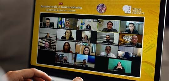 Imagem de um notebook exibindo a tela do evento on-line. Na live, o fundo é amarelo e na parte s...