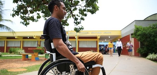 Fotografia de um homem, que veste camisa preta e calça laranja, sentado em uma cadeira de rodas....