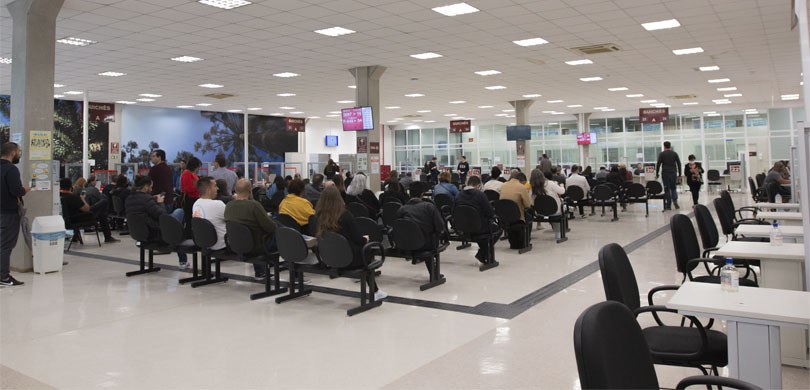 Fotografia de uma grande sala, com várias cadeiras ao centro, onde pessoas estão sentadas aguard...