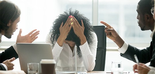 Foto de três pessoas. No centro, há uma mulher sentada à mesa com a cabeça abaixada e com as dua...