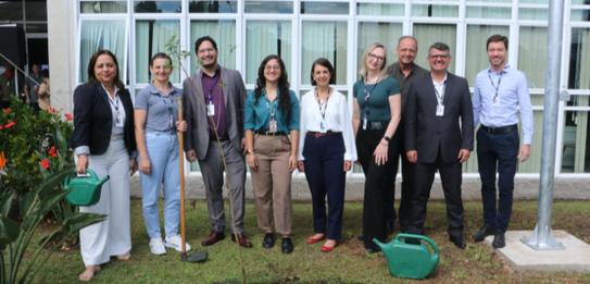 Fotografia de nove pessoas, entre homens e mulheres, posando sorrindo em frente a um edifício co...