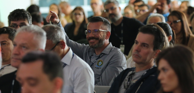 Fotografia na qual é possível ver um servidor sorrindo e apontando a mão direita para o palco. O...