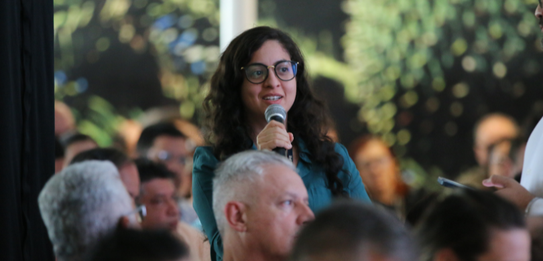Fotografia de nove pessoas, entre homens e mulheres, posando sorrindo em frente a um edifício co...