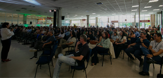 A Diretora-Geral Solange Maria Vieira fala para a plateia segurando um microfone. Atrás dela, um...