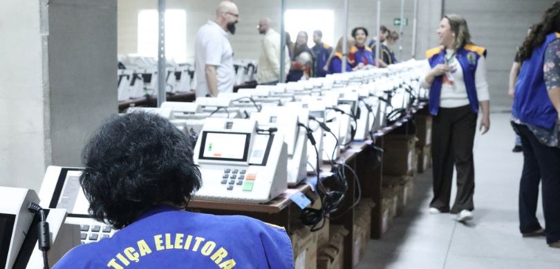 Fotografia em galpão amplo na qual diversas pessoas trabalham na preparação de urnas eletrônicas...