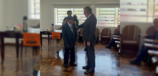 Fotografia de um homem de terno e gravata parado ao lado de uma cabina de votação em uma sala de...