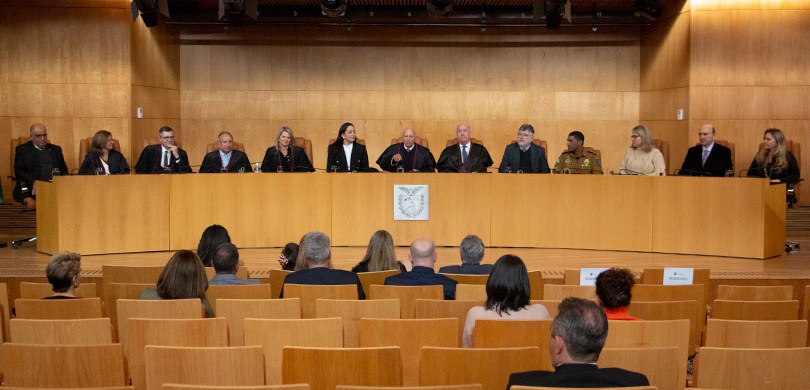 Fotografia de uma mesa de autoridades com 13 pessoas, entre homens e mulheres, diante de uma pla...