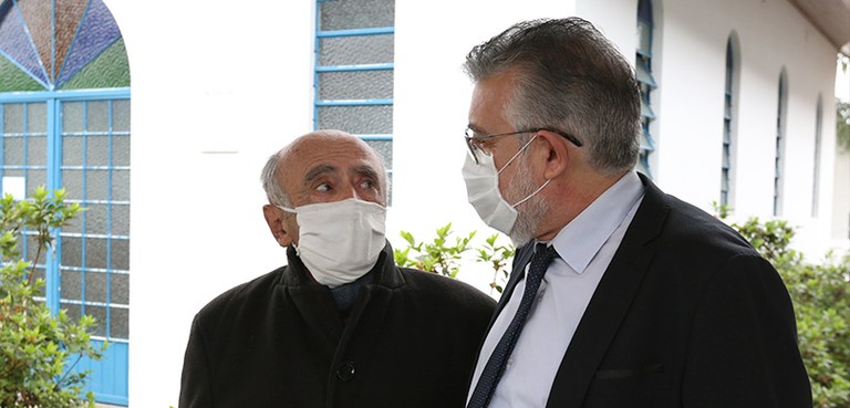 Dois homens se olham. Ao fundo, um imovel com janelas e portas de vidro. Os dois usam máscara.