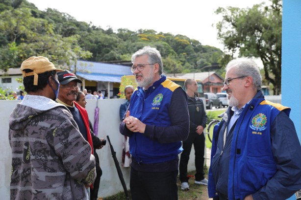 Fotografia de duas pessoas com o colete azul da Justiça Eleitoral conversando com moradores em u...