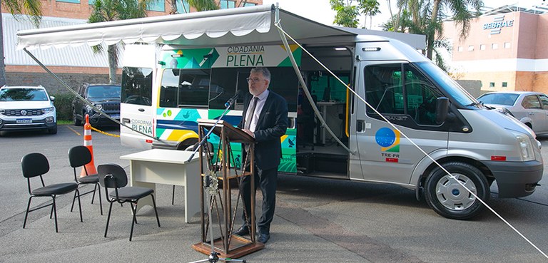 Fotografia de um homem de terno falando em um púlpito na frente de uma van prata, em que se lê: ...