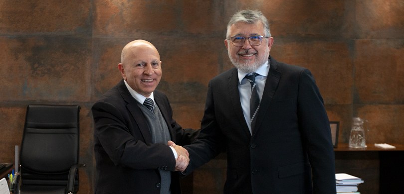 Dois homens apertam as mãos e sorriem. Os dois usam terno. Ao fundo, uma parede marrom.