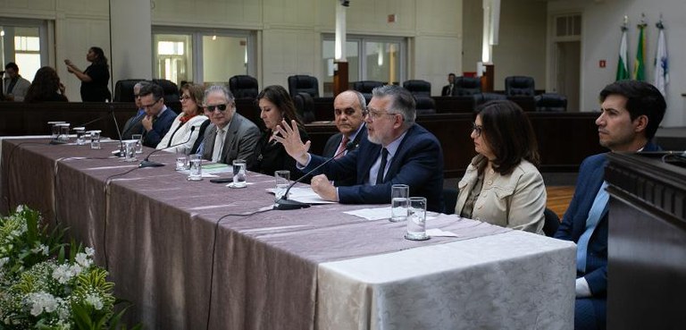 Uma mesa decorada com uma toalha roxa com alguns copos e microfones em cima. Há nove autoridades...