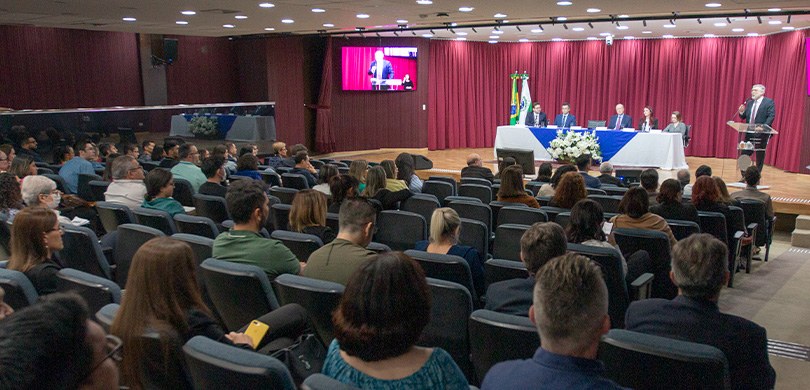 Fotografia de várias pessoas sentadas em cadeiras em um auditório. Ao fundo, uma mesa com cinco ...