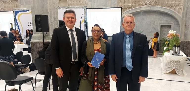 Presidente do TRE-PR e diretor-executivo da EJE-PR recebem homenagem no XXII Codeje Fotografia de dois homens de terno e uma mulher, ao centro, que usa um vestido estampado, blazer...