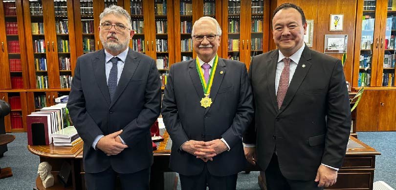 Fotografia de quatro homens de terno sorrindo em uma sala. O homem ao centro usa no pescoço uma ...