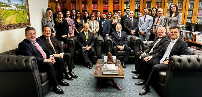 Fotografia de seis homens de terno sentados em poltronas pretas em um meio círculo. Ao fundo, ou...