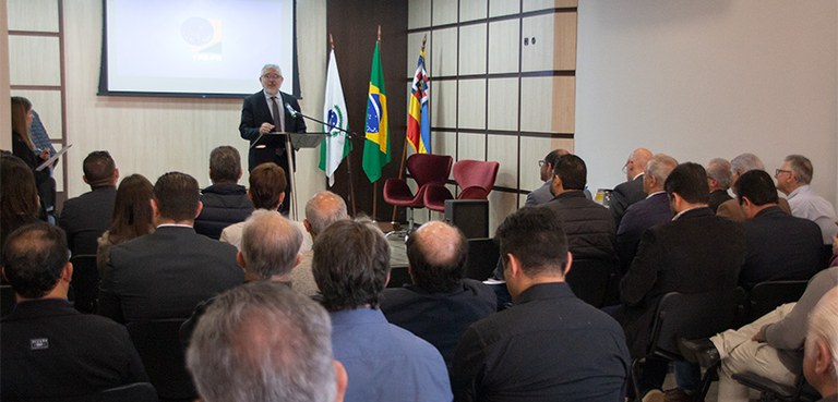 Fotografia de um pequeno auditório. À frente, discursa o presidente do TRE-PR sob o palco. À dir...