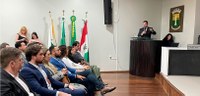 Fotografia em que se observa um auditório ou plenário com diversas pessoas assistindo a uma apre...