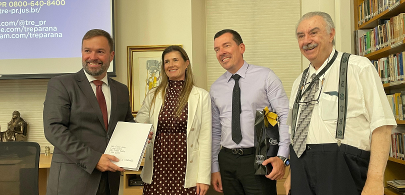 Fotografia colorida em plano médio que retrata quatro pessoas, três homens e uma mulher, em pé e...