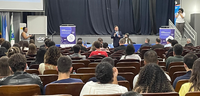 Fotografia de um auditório. Em primeiro plano e no plano médio, observa-se a parte posterior das...