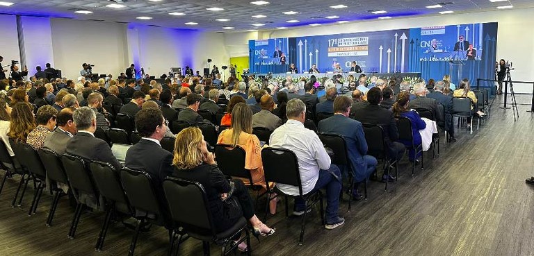 Foto do fundo de um auditório, no qual se observam várias pessoas sentadas de costas e, bem no f...