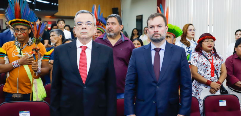 Fotografia de diversas pessoas em pé, em um ambiente interno. Em primeiro plano, dois homens, en...