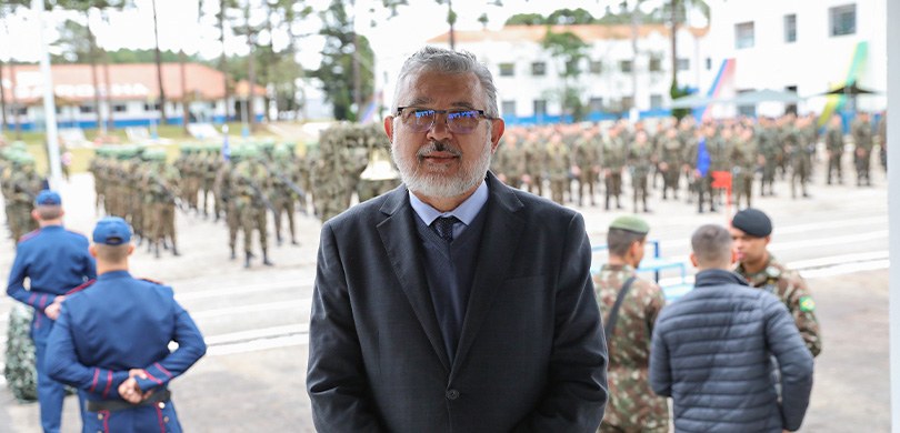 Um homem está parado, vestindo terno, a frente. Atrás está um batalhão do exército, com várias p...