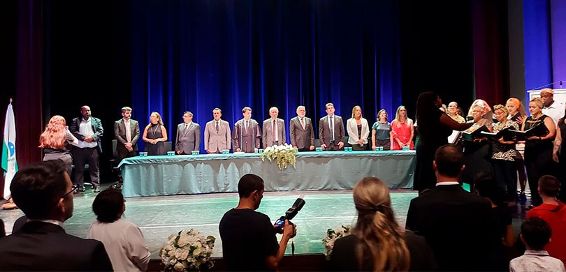 Fotografia da mesa de autoridades em um palco. Ao fundo uma cortina azul, ao lado um coral e uma...