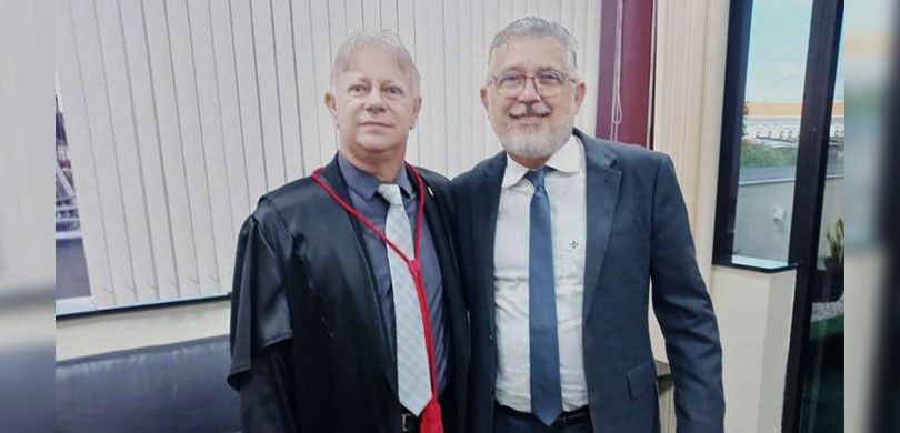 Dois homens posam sorrindo para a foto. Um deles está usando terno e outro de toga.