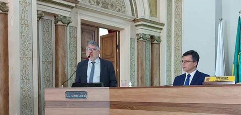 À esquerda, um homem de óculos, vestindo terno escuro e camisa branca, discursa na mesa do plená...