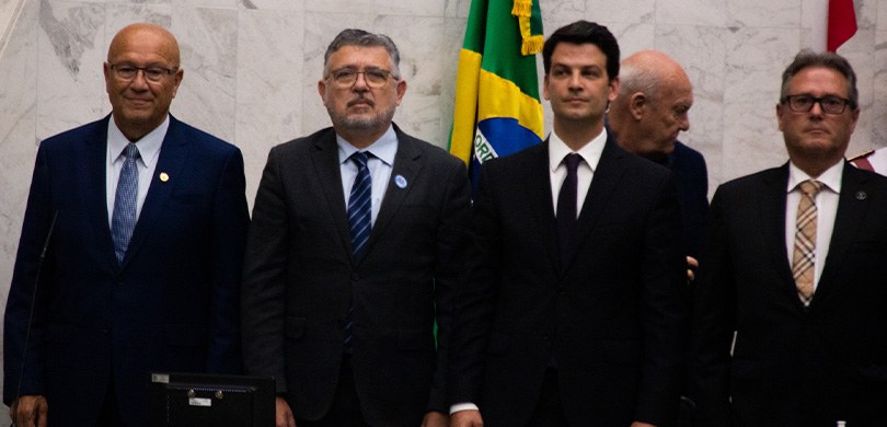 Quatro homens de terno posam para a foto. Ao fundo, um homem e a bandeira do Brasil.