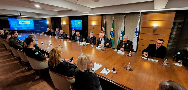 Fotografia de uma comprida mesa de reuniões. Há diversas pessoas sentadas frente a frente e papé...