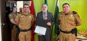 Três homens posam para a foto em uma sala com parede verde ao fundo e três bandeiras. Ao centro,...