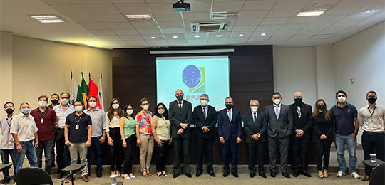 Fotografia de um grupo de pessoas em pé, lado a lado. Ao fundo, no centro do salão, há um banner...