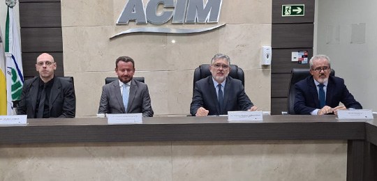 Fotografia de quatro homens sentados atrás de uma mesa. Acima, aparece a sigla ACIM em um painel...