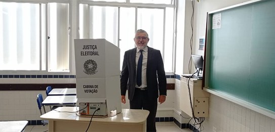 Fotografia de um homem de terno e gravata parado ao lado de uma cabina de votação em uma sala de...